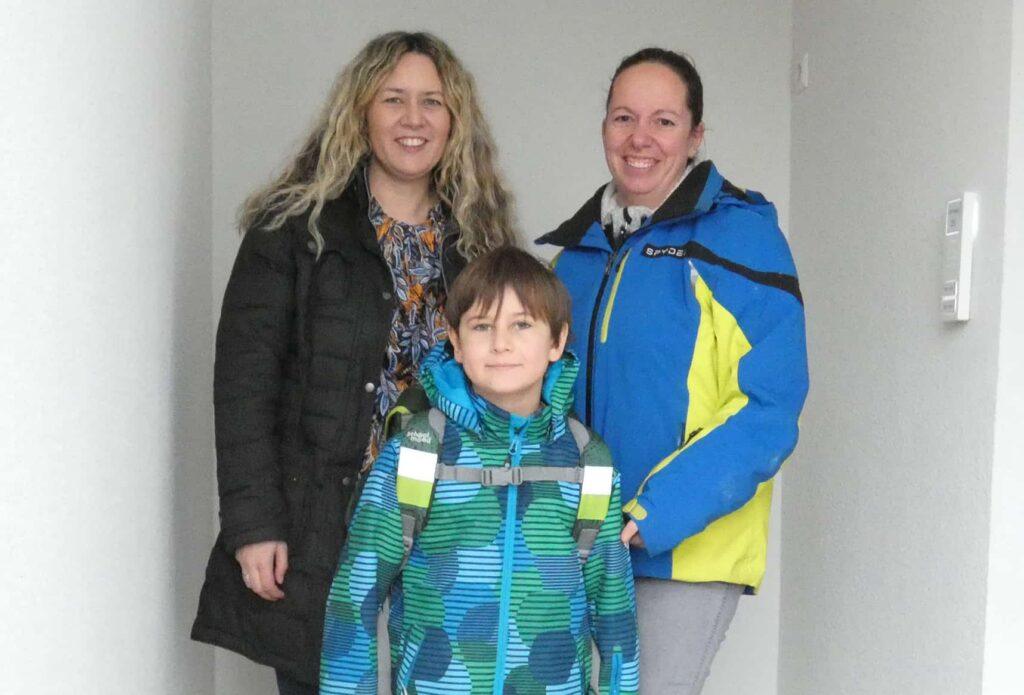 Zwei Frauen stehen in einem Haus hinter einem Jungen, der eine grün-blau gemusterte Jacke und einen Rucksack trägt. Alle drei schauen in die Kamera und lächeln.