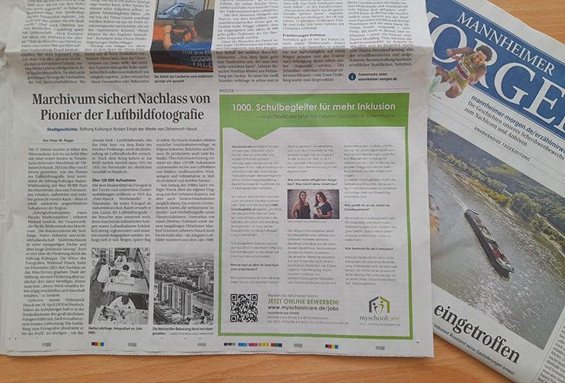 Auf einem Tisch liegt eine aufgeschlagene deutsche Zeitung mit einem Artikel über Luftaufnahmen und einer Anzeige zur Inklusion in Schulen; daneben liegt eine weitere gefaltete Zeitung mit dem Titel "Mannheimer Morgen", die auf der Titelseite eine Flussszene zeigt.