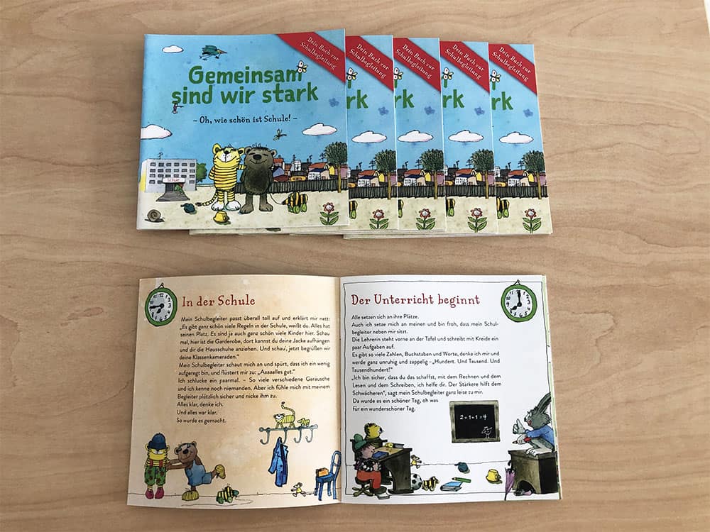 Fünf Exemplare eines deutschen Kinderbuchs mit dem Titel Gemeinsam sind wir stark liegen gestapelt auf einer Holzfläche. Darunter ist ein Buch aufgeschlagen und zeigt illustrierte Seiten und einen deutschen Text über die Schule.