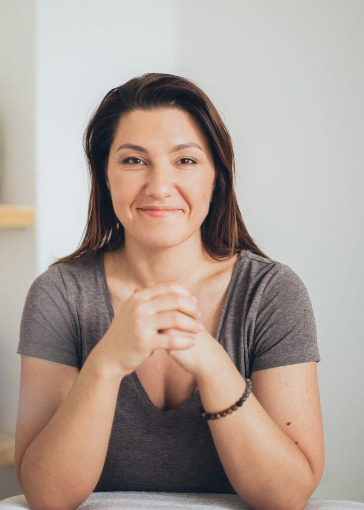 Eine Frau mit langen braunen Haaren sitzt an einem Tisch, faltet ihre Hände und lächelt sanft. Sie trägt ein graues, kurzärmeliges Hemd und ein Perlenarmband. Der Hintergrund ist neutral.