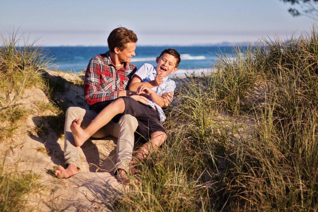 Ein Mann und ein Junge sitzen barfuß auf sandigem Boden zwischen hohem Gras am Strand. Der Mann kitzelt den Jungen, der lacht. Im Hintergrund sind das Meer und der Himmel zu sehen.