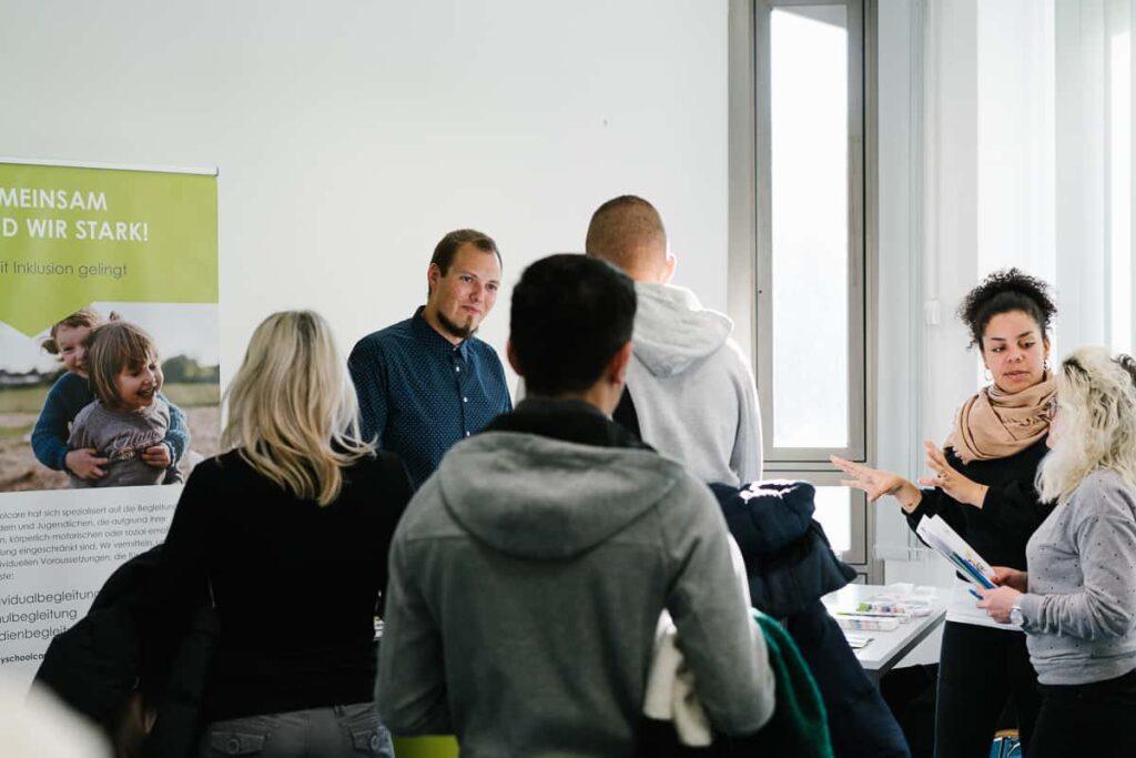 Eine Gruppe von Menschen steht in einem hell erleuchteten Raum um einen Tisch herum und unterhält sich. Ein Mann steht hinter dem Tisch, während eine Frau mit ihren Händen gestikuliert. Im Hintergrund sind eine Fahne und Fenster zu sehen.
