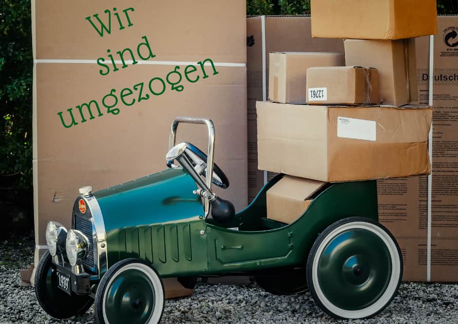 Ein grünes Spielzeugauto ist neben Pappkartons geparkt, auf dessen Sitz sich weitere Kartons stapeln. Im Hintergrund stehen große Pappkartons. Auf der linken Seite ist der deutsche Text "Wir sind umgezogen" zu lesen.