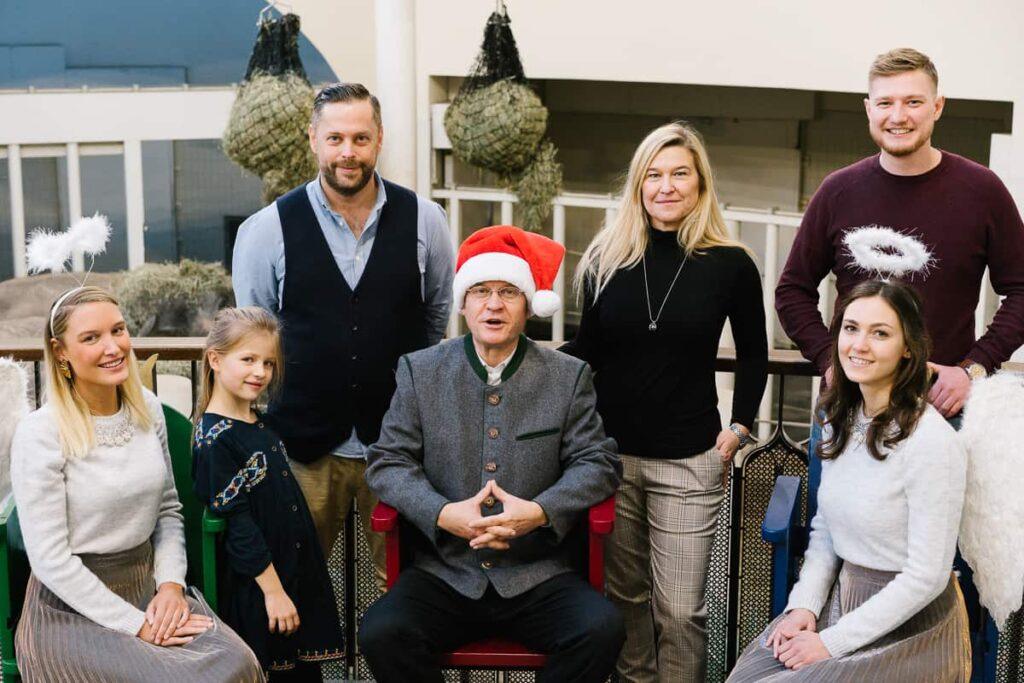 Eine Gruppe von sechs Erwachsenen und einem Kind posiert in einem Haus. Der Mann in der Mitte trägt eine Weihnachtsmannmütze und eine Anzugsjacke. Zwei Frauen tragen Engelskränze. Sie alle stehen oder sitzen vor einem Geländer, mit großen grünen Kränzen im Hintergrund.