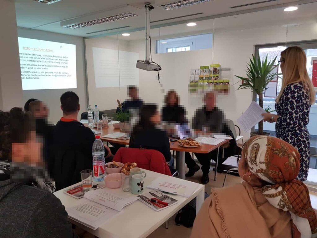 Eine Gruppe von Personen sitzt um einen Tisch in einem Besprechungsraum und hört einer Frau zu, die Informationen auf einen Bildschirm projiziert. Auf dem Tisch liegen Unterlagen, Becher und Snacks.