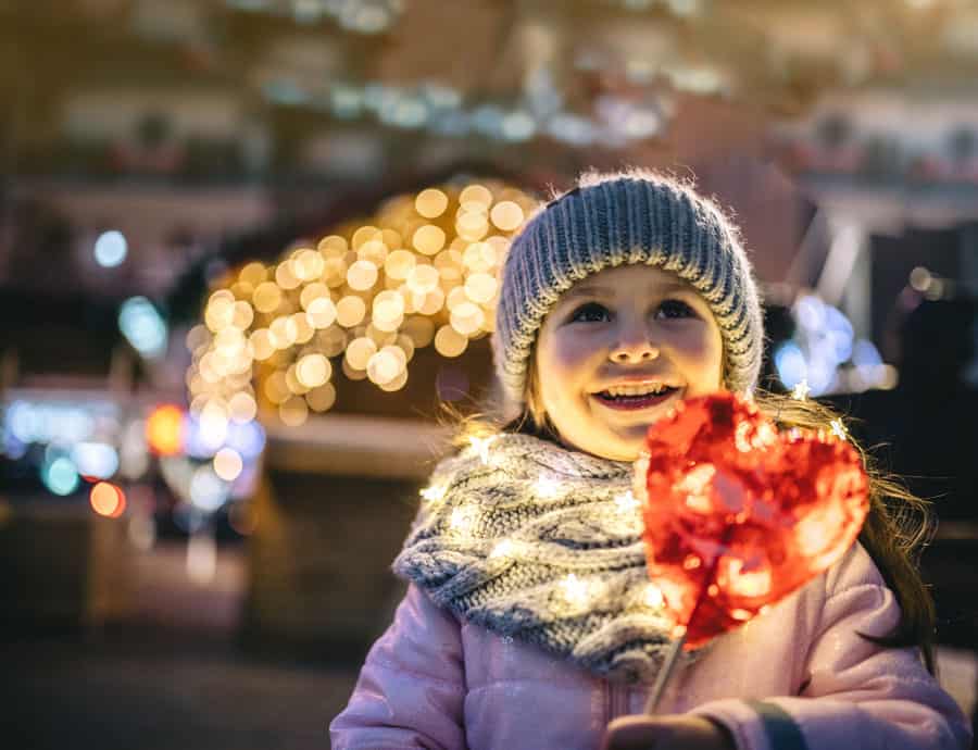 Ein kleines Kind mit Strickmütze und Schal steht nachts im Freien, lächelt und hält einen herzförmigen roten Lutscher in der Hand. Lichterketten sind um das Kind gewickelt, im Hintergrund sind verschwommene Lichterketten zu sehen.