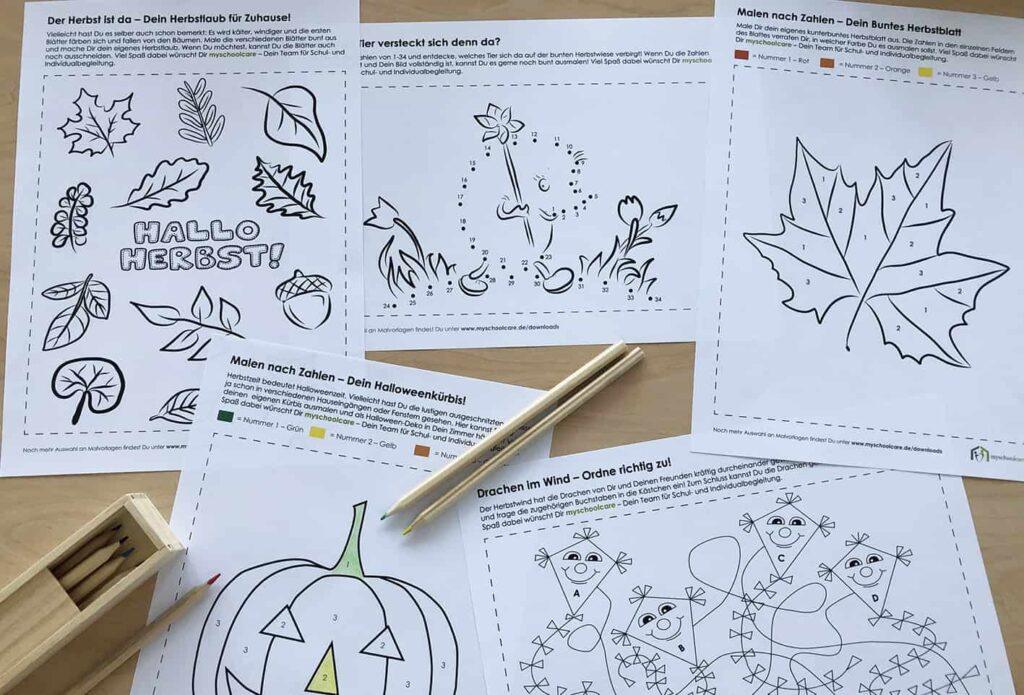 Fünf Malvorlagen zum Thema Herbst liegen auf einem Tisch mit drei Buntstiften und einer hölzernen Stiftebox. Auf den Seiten sind Blätter, ein Kürbis, Pilze und andere herbstliche Motive abgebildet, die teilweise auf Deutsch beschriftet sind.