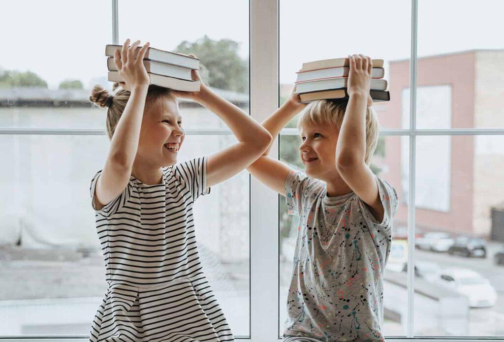 Zwei Kinder sitzen an einem Fenster und balancieren jeweils einen Stapel Bücher auf dem Kopf. Sie lächeln und schauen sich gegenseitig an. Ein Kind trägt ein gestreiftes Kleid, das andere ein hell gemustertes Hemd.