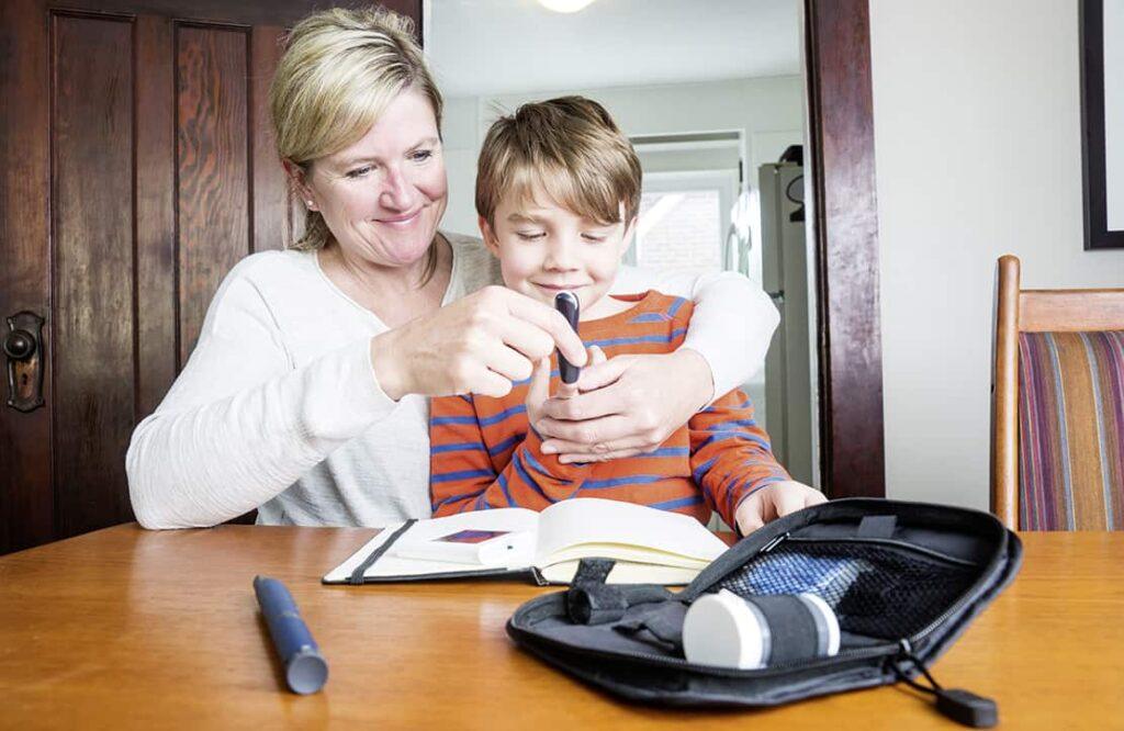 Eine Frau hilft einem Jungen an einem Tisch, ein Blutzuckermessgerät zu bedienen. Vor ihnen auf dem Tisch liegen Diabetes-Zubehör und ein aufgeschlagenes Notizbuch. Beide scheinen auf ihre Aufgabe konzentriert zu sein.