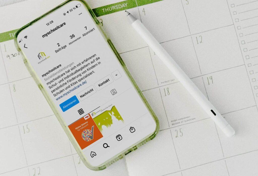 Ein Smartphone mit einem geöffneten Instagram-Profil liegt auf einem Kalender; ein weißer Stift ist in der Nähe platziert. Das Instagram-Profil zeigt ein Profilbild, Beiträge und Follower-Details. Die Kalendertage sind teilweise sichtbar.