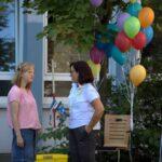 Zwei Frauen unterhalten sich im Freien in der Nähe eines Baumes, an dem bunte Luftballons befestigt sind. Eine Frau trägt ein rosa Hemd und einen Jeansrock, die andere ein weißes Hemd und hält eine Wasserflasche in der Hand. Im Hintergrund sind ein Stuhl und Reinigungsgeräte zu sehen.