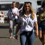 Eine Frau in weißem Polohemd und Jeans geht lächelnd im Freien spazieren und trägt ein Schlüsselband um den Hals. Im Hintergrund sind andere Menschen und ein Wohnmobil zu sehen. Es ist sonnig.