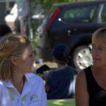 Zwei Frauen sitzen im Freien und unterhalten sich. Die eine trägt ein weißes Poloshirt mit grünem Logo, die andere ein ärmelloses schwarzes Oberteil. Im Hintergrund sind ein Kind und ein Auto zu sehen.