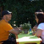 Ein Mann in einem orangefarbenen Hemd und eine Frau in einem weißen Hemd sitzen an einem Tisch mit einer grünen Tischdecke im Freien und unterhalten sich. Eine kleine Pflanze und eine blaue Glasflasche stehen auf dem Tisch. Im Hintergrund ist grünes Laub zu sehen.