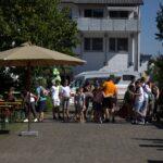 Eine Gruppe von Menschen versammelt sich im Freien bei Picknicktischen und einem Sonnenschirm. Im Hintergrund ist ein weißer Lieferwagen geparkt. Ein Schild und grüne Luftballons sind zu sehen, und im Hintergrund sind Wohngebäude zu sehen.