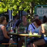 Eine Gruppe von Menschen sitzt um einen Holztisch im Freien, unterhält sich und isst. Auf dem Tisch stehen Getränke und Schüsseln. Im Hintergrund ist ein grünes Schild mit einem Kreuz und dem Wort "Jugend" zu sehen. Bäume spenden Schatten.