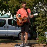 Eine Person in einem korallenroten T-Shirt und schwarzen Shorts spielt eine akustische Gitarre und singt in ein Mikrofon auf einer Bühne im Freien. Im Hintergrund sind ein silberner Lieferwagen und grünes Laub zu sehen.