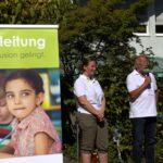 Ein Mann, der in ein Mikrofon spricht, steht neben einer Frau im Freien, beide tragen weiße Hemden. Auf der linken Seite des Bildes ist ein Banner mit dem Foto eines jungen Mädchens und deutschem Text zu sehen. Im Hintergrund sind Bäume und ein Gebäude zu sehen.