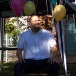 Ein Mann mit Bart sitzt auf einem Stuhl im Freien unter einem Vordach, umgeben von bunten Luftballons. Er trägt ein weißes Polohemd mit einem Logo und einem Namensschild, lächelt und schaut leicht nach links.