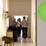 Eine Gruppe von Personen steht in einem Flur in der Nähe einer offenen Tür. Auf der linken Seite befinden sich Stühle, ein Drucker und ein Regal mit einer Pflanze. Blaue Luftballons sind teilweise im Raum hinter der Tür zu sehen.