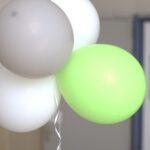 Ein Bündel von vier Luftballons, drei weiße und ein hellgrüner, ist mit einem weißen Band vor einem unscharfen Innenhintergrund zusammengebunden.