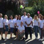 Eine Gruppe von elf Personen, die alle weiße Polohemden und Namensschilder tragen, posieren im Freien im Sonnenlicht. Einige stehen, einer kniet. Im Hintergrund sind grüne Bäume, Luftballons und andere Personen unscharf zu erkennen.