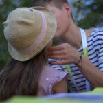 Ein Erwachsener in einem gestreiften Hemd berührt sanft die Wange eines Kindes, das einen Strohhut und ein rosa Kleid trägt. Die Szene scheint im Freien zu spielen, mit Grünzeug im Hintergrund.