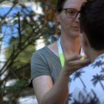 Eine Frau mit Brille schminkt eine Person, die vor ihr steht. Sie hat ein Schlüsselband um den Hals und steht im Freien, wobei im Hintergrund Grünflächen zu sehen sind.