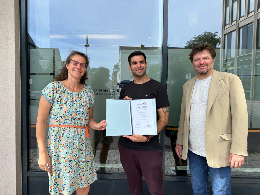 Drei Erwachsene stehen vor einer Glastür. Die Person in der Mitte hält eine offene Mappe mit einer Urkunde in der Hand. Die beiden Personen auf beiden Seiten lächeln. Im Hintergrund sind einige Gebäude und eine Spiegelung eines Baumes zu sehen.
